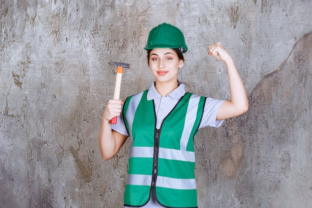 Ingenieurin in grünem helm, die eine holzaxt für reparaturarbeiten hält und ihre armmuskulatur zeigt