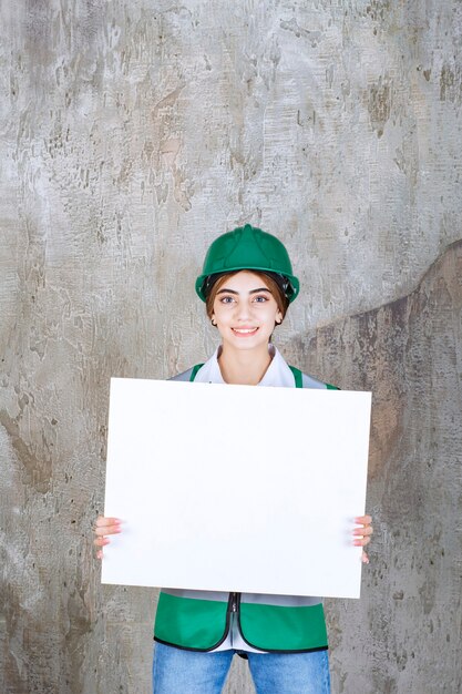 Ingenieurin in grüner Uniform und Helm, die eine rechteckige Infotafel hält.