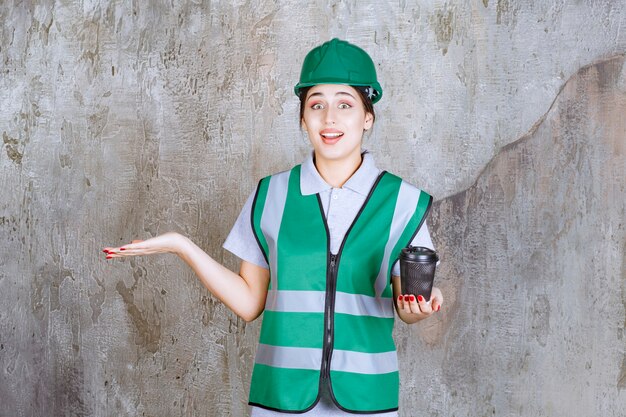 Ingenieurin in grüner Uniform und Helm, die eine schwarze Kaffeetasse hält und etwas beiseite zeigt.