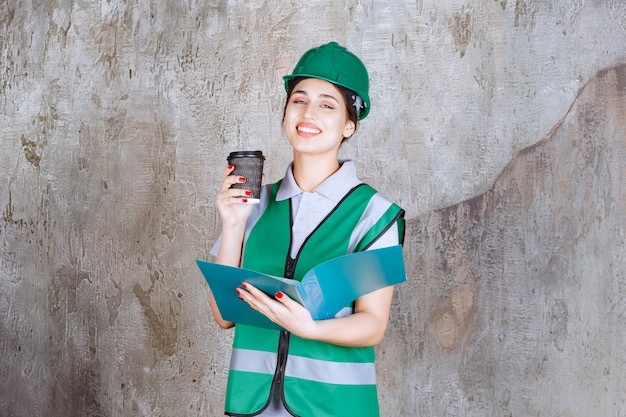 Ingenieurin in grüner Uniform und Helm mit schwarzer Kaffeetasse und blauem Projektordner