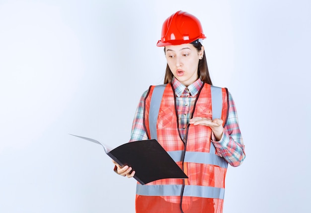 Ingenieurin mit rotem helm, die einen schwarzen projektplan hält und verwirrt aussieht.