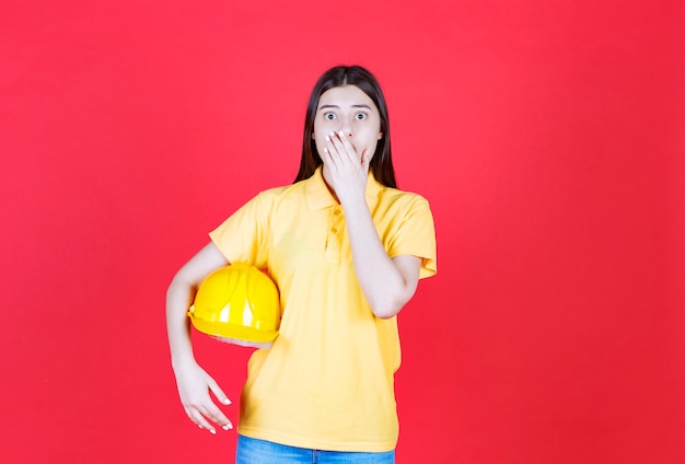 Ingenieursmädchen im gelben Dresscode, das einen gelben Schutzhelm hält und erschrocken und verängstigt aussieht.