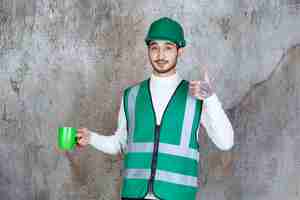 Kostenloses Foto ingenieursmann in gelber uniform und helm, der eine grüne kaffeetasse hält und das produkt genießt.