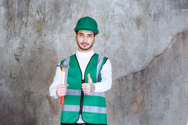 Ingenieursmann in gelber Uniform und Helm, der eine Holzaxt zur Reparatur hält und Freudenhandzeichen zeigt