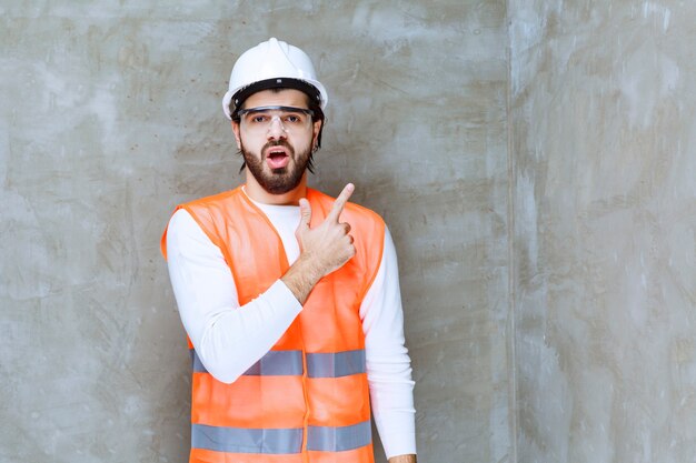 Ingenieursmann mit weißem Helm und Schutzbrille, der auf etwas zeigt.