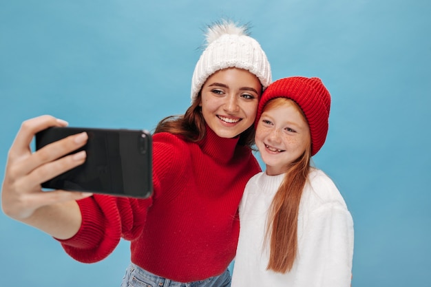 Ingwer kleines Mädchen in rotem Hut und leichtem weiten Pullover posiert und macht Foto mit ihrer charmanten Schwester in weißer Mütze und cooler Kleidung