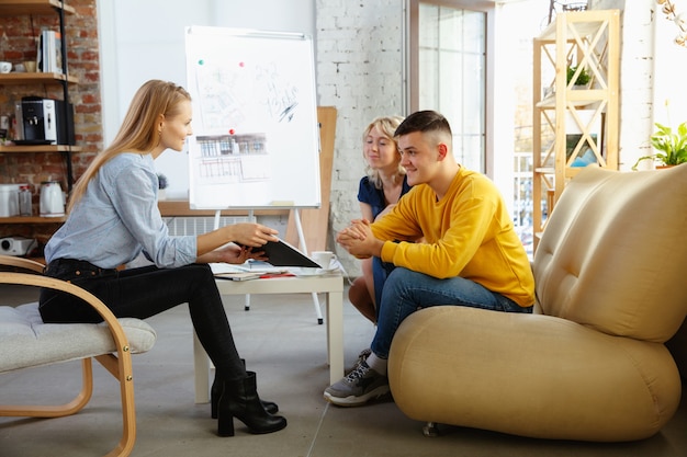 Innenarchitekt, der mit jungem Paar arbeitet. Schöne Familie und professioneller Designer oder Architekt, der das Konzept des zukünftigen Interieurs bespricht und mit Farbpalette, Raumzeichnungen im modernen Büro arbeitet.