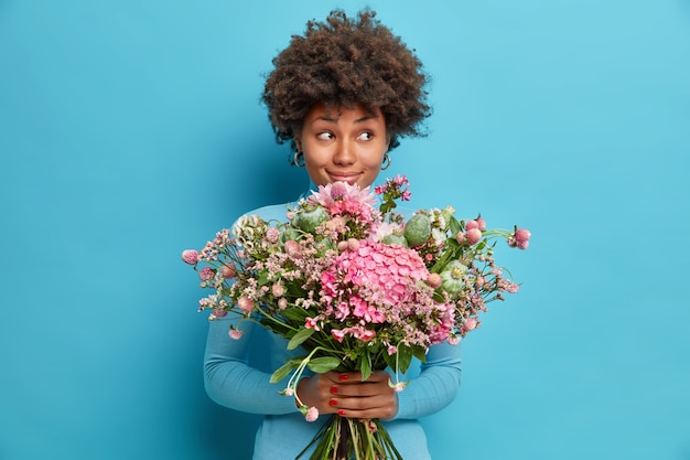 Innenaufnahme der afroamerikanischen Frau erhält schöne Blumen genießt romantische Verabredungsblicke mit verträumtem nachdenklichem Ausdruck beiseite erhält Blumenstrauß vom geheimen Bewunderer oder Liebhaber isoliert auf blauer Studiowand