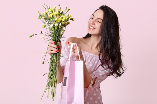 Innenaufnahme der angenehm aussehenden jungen Frau lächelt sanft, modisches Kleid gekleidet, hält Geschenktüte und Blumen, posiert auf hellrosa, erfreut. Frauentagskonzept