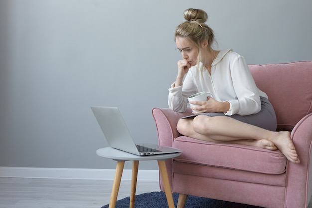 Innenaufnahme der attraktiven jungen Frau mit Haarknoten, die barfuß im Sessel mit Tasse Kaffee sitzt und Webinar über Laptop-Computer betrachtet, online lernt, konzentrierten Blick konzentriert