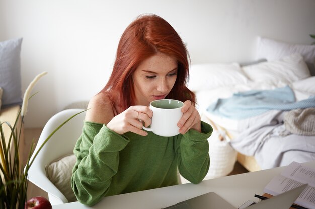 Innenaufnahme der attraktiven jungen rothaarigen Frau in der Freizeitkleidung beim Morgenkaffee, am Tisch in ihrem Zimmer sitzend