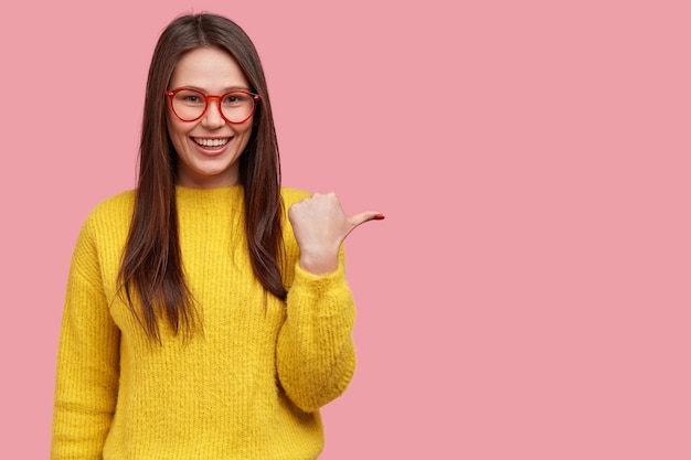 Innenaufnahme der attraktiven lächelnden Frau zeigt beiseite und lächelt zufrieden, zeigt kühlen Kopierraum, trägt optische Brille, gelbe Kleidung