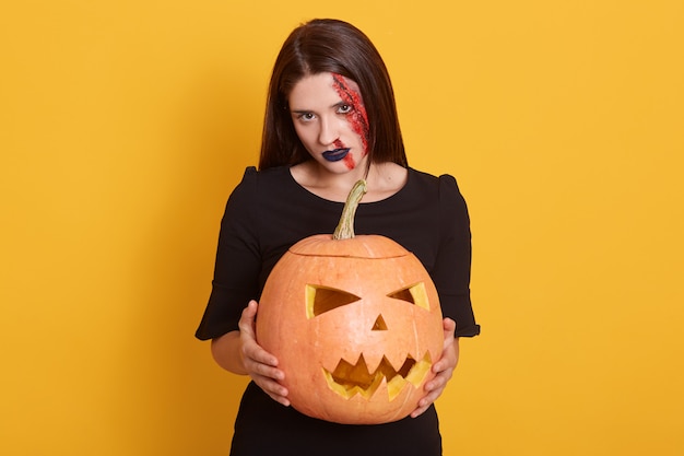 Innenaufnahme der brünetten Frau im Halloween-Make-up, der Kürbis über gelb hält