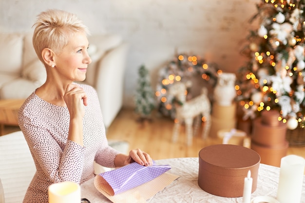 Innenaufnahme der fröhlichen reifen kurzhaarigen europäischen Frau, die sich auf Neujahrs- oder Weihnachtsfeier vorbereitet, im Wohnzimmer mit Geschenkpapier auf Tisch sitzend, nachdenklich nachdenklich aussehend, lächelnd