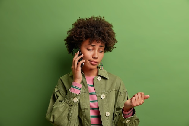 Innenaufnahme der gelangweilten Afroamerikanerin hat Telefongespräch, schaut auf ihre neue Maniküre, verärgert Ausdruck, gut gekleidet, steht