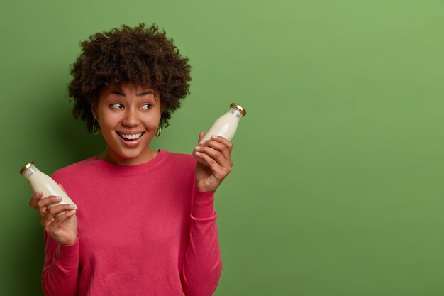 Innenaufnahme der glücklichen Frau trinkt Bio-Mandelmilch, posiert mit gesundem vegetarischem Produkt in der Flasche, hat gesunden Lebensstil, schaut mit Lächeln beiseite, trägt rosigen Pullover, lokalisiert auf grüner Wand