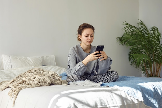 Innenaufnahme der lässig gekleideten attraktiven jungen Frau, die mit gekreuzten Beinen auf einem ungelösten Bett sitzt