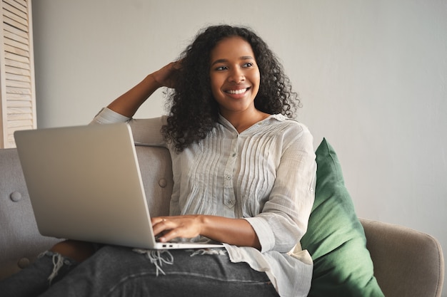 Innenaufnahme der positiven charmanten jungen Afroamerikanerin gekleidet in stilvolle Kleidung, die auf Sofa mit tragbarem Computer auf ihrem Schoß entspannt, online einkauft, mit niedlichem fröhlichem Lächeln wegschauend