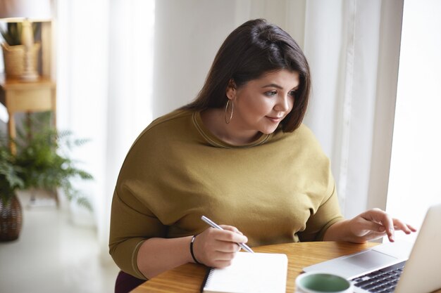 Innenaufnahme der übergewichtigen Übergröße schöne junge brünette Dame in der stilvollen Kleidung, die am Schreibtisch mit offenem Laptop, Tasse Kaffee sitzt und Informationen in ihrem Tagebuch aufschreibt, online lernt