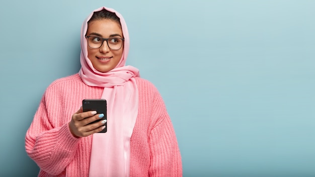 Innenaufnahme der verträumten positiven jungen Frau benutzt Handy, prüft Nachrichten, gekleidet in rosa gestrickten Pullover, isoliert über blauer Wand mit Kopienraum für Ihre Werbung, liest Artikel