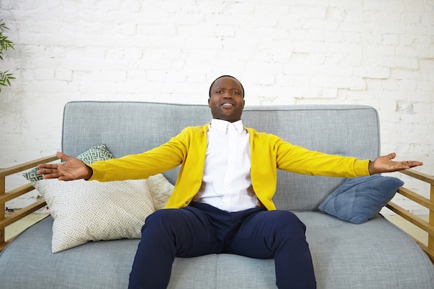 Innenaufnahme des emotionalen fröhlichen jungen Afroamerikaners in der stilvollen Kleidung, die auf bequemem grauem Sofa im Wohnzimmer sitzt, Arme auseinander hält, aufgeregten Gesichtsausdruck überglücklich