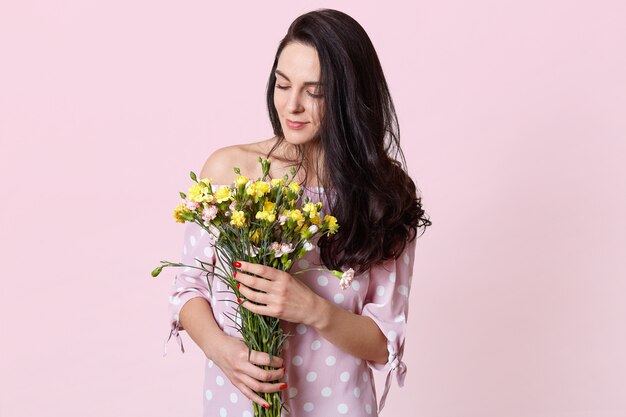 Innenaufnahme des erfreuten dunkelhaarigen weiblichen Modells hält Blumenstrauß, gekleidet in modischem Kleid, lokalisiert auf Rosa. Romantische attraktive Frau erhält Blumen am 8. März