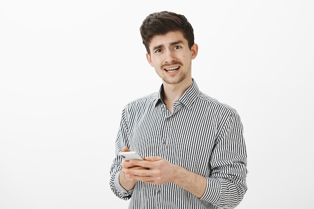 Innenaufnahme des frustrierten unzufriedenen reifen Mannes mit Schnurrbart im lässigen gestreiften Hemd, der befragt schaut, während er Frage stellt und Smartphone hält, verwirrende Nachricht empfängt