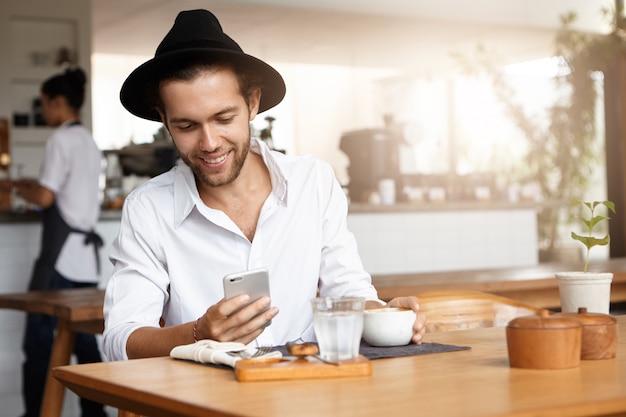 Innenaufnahme des hübschen jungen Mannes, der Hut und weißes Hemd trägt, die glücklich lächeln, während sie SMS auf Handy lesen, seine Freundin online mit kostenlosem Wi-Fi während des Mittagessens im Café mitteilend
