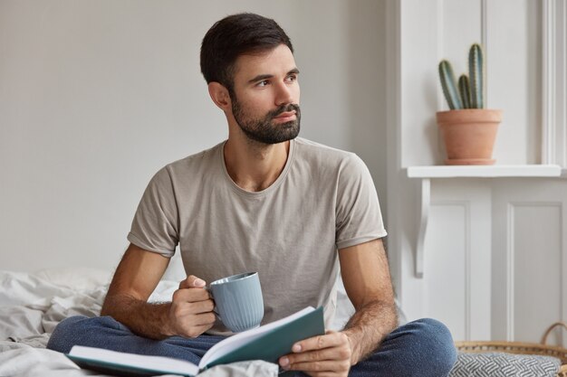 Innenaufnahme des nachdenklichen jungen kaukasischen Kerls mit dickem Bart, der tief in Gedanken ist, Buch und Tasse Tee hält, posiert auf Bett