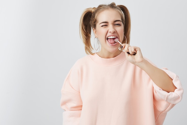Innenaufnahme des positiven Teenager-Mädchens mit zwei Pferdeschwänzen, die rosa Sweatshirt tragen, lokalisiert auf grauem Studiohintergrund mit Lutscher in ihrem Mund. Blondes lustiges Mädchen, das Lutscher isst.