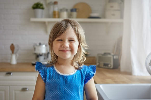 Innenaufnahme des schönen lustigen kleinen Mädchens mit hellem Haar und weicher Haut, die freudig lächelnd, positive Gefühle und Freude ausdrückend, niedliches blaues Kleid tragend. Kindheit, Glück und Unschuld