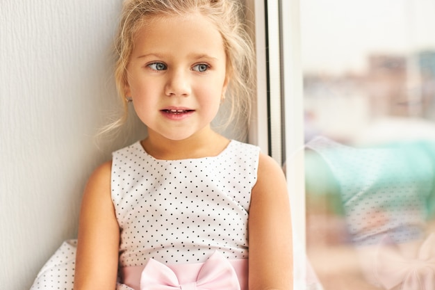 Innenaufnahme des schönen traurigen kaukasischen Mädchens im gepunkteten Kleid, das auf Fensterbank sitzt, verärgertes Aussehen hat, sich einsam fühlt und auf Eltern von der Arbeit wartet. Menschen, Kinder, Lebensstil und Einsamkeit Konzept