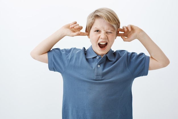 Kostenloses Foto innenaufnahme des unglücklichen verärgerten jungen europäischen kindes im blauen t-shirt, schreiend oder schreiend, ohren mit zeigefingern bedeckend, mit einem auge spähend