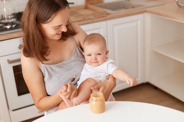 Innenaufnahme einer dunkelhaarigen Frau füttert ihre kleine Tochter mit Obst- oder Gemüsepüree, Mutter schaut ihr süßes Baby mit Liebe an, gesunde Fütterung.