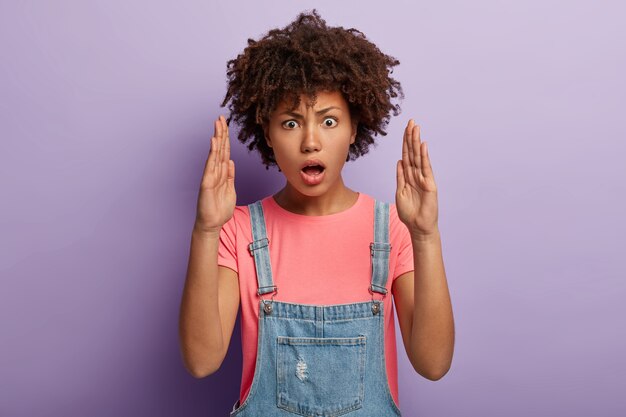 Innenaufnahme einer emotional überraschten jungen Frau mit einem Afro, der in Overalls posiert