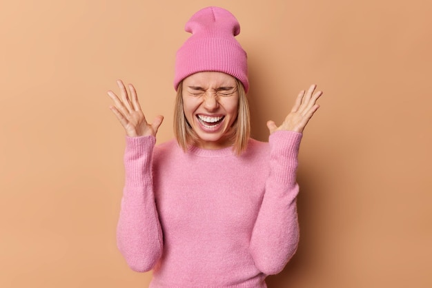 Kostenloses Foto innenaufnahme einer emotionalen jungen frau hält die handflächen erhoben, schreit laut die stirn, das gesicht zeigt weiße zähne, reagiert auf gute unerwartete nachrichten, gekleidet in rosa rollkragenpullover und hut, isoliert auf brauner wand