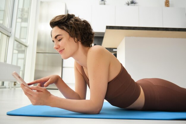 Innenaufnahme einer fitten und gesunden Frau, die zu Hause auf einer Yogamatte trainiert und dabei ihr Smartphone benutzt