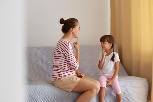 Innenaufnahme einer Frau mit Haarverbot, die mit einem kleinen Mädchen auf dem Sofa sitzt und dem Kind zeigt, wie man Geräusche ausspricht, Privatunterricht mit einem professionellen Sprachpathologen.