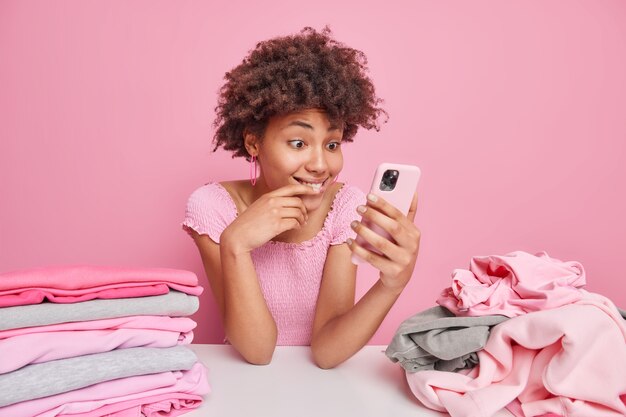 Innenaufnahme einer fröhlichen Frau mit Afro-Haar macht Pause, nachdem sie Hausarbeit gemacht hat und Kleidung faltet überprüft Newsfeed im Smartphone sitzt am Tisch isoliert über Pink
