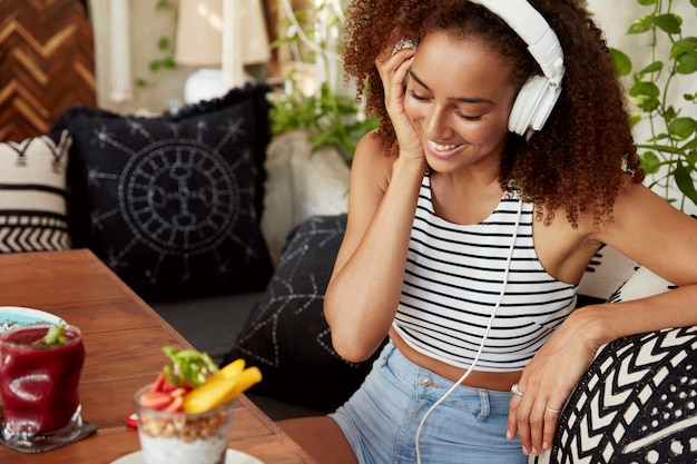 Innenaufnahme einer glücklichen Frau mit dunkler Haut und Afro-Frisur hört Audiospur in Kopfhörern, sieht im Handy positiv aus, ruht während der Pause, verwendet moderne Technologien, ist mit dem Internet verbunden