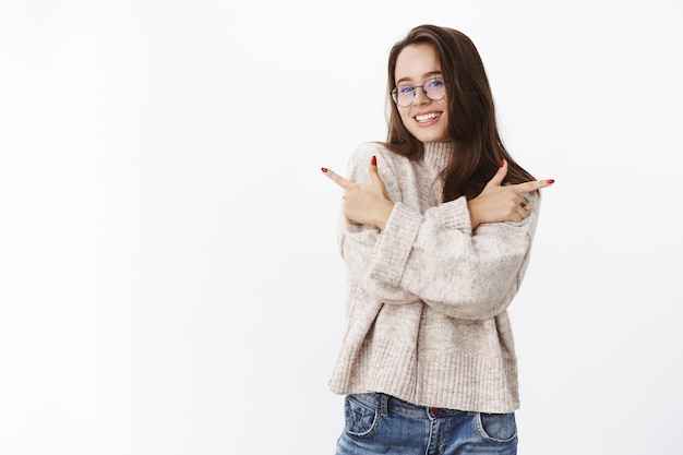 Innenaufnahme einer glücklichen hübschen Brünette in Brille und warmem Pullover, die die Arme über der Brust kreuzt und nach links und rechts zeigt