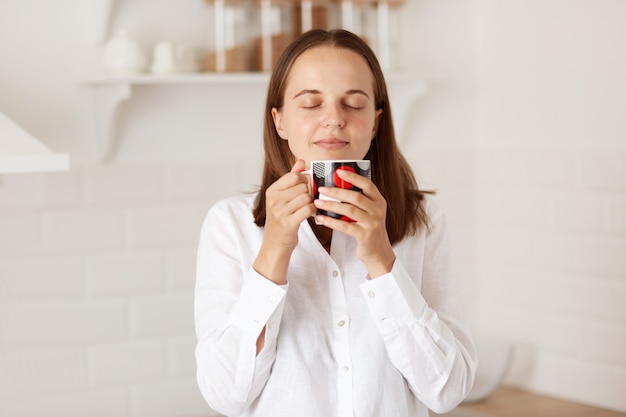 Innenaufnahme einer glücklichen hübschen Frau, die morgens eine Tasse Kaffee oder Tee in der Küche genießt, heiße Getränke trinkt und mit geschlossenen Augen mit entspanntem Ausdruck steht.