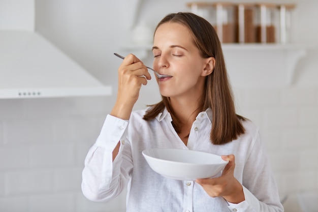 Innenaufnahme einer glücklichen jungen Frau, die zu Hause Suppe isst. Genießen Sie ein leckeres Frühstück oder Abendessen, tragen Sie ein weißes Hemd und posieren Sie mit einer hellen Küche im Hintergrund.