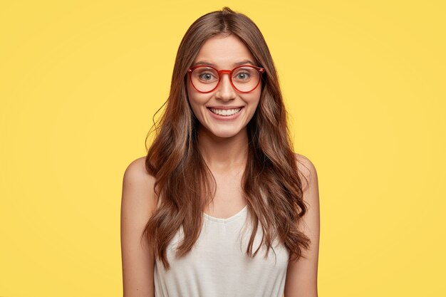 Innenaufnahme einer gut aussehenden jungen Brünette mit Brille, die gegen die gelbe Wand aufwirft