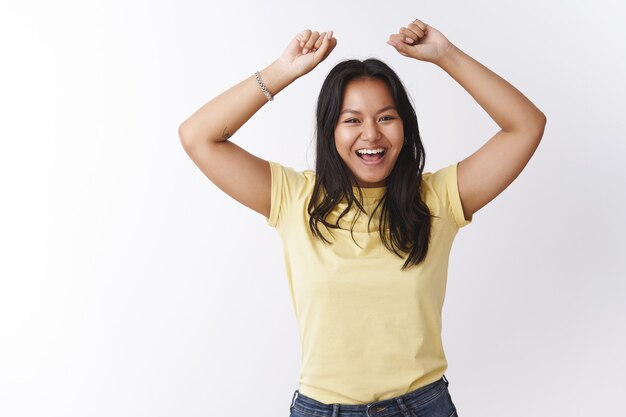 Innenaufnahme einer optimistisch glücklichen und emotionalen jungen malaysischen Frau, die spielerisch springt und lacht, in die Kamera grinst und die Hände hebt, die glücklich tanzen und Spaß haben, gute Laune über weiße Wand zu haben