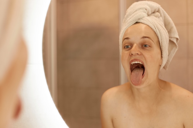 Innenaufnahme einer sonnigen Frau, die nach dem Duschen im Badezimmer posiert, ihr Spiegelbild mit herausgestreckter Zunge betrachtet, ihre Zunge betrachtet, hygienische Verfahren.