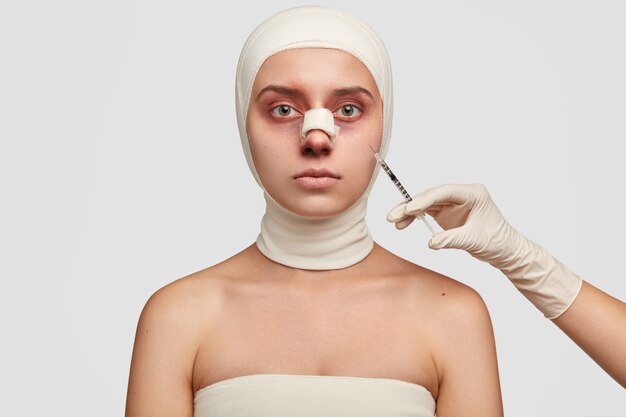 Innenaufnahme einer verletzten Frau mit blasser Haut, hat Gips auf der Nase, ist mit Bandagen umwickelt, erhält eine Injektion vom Chirurgen und hat einen ernsthaften Gesichtsausdruck.