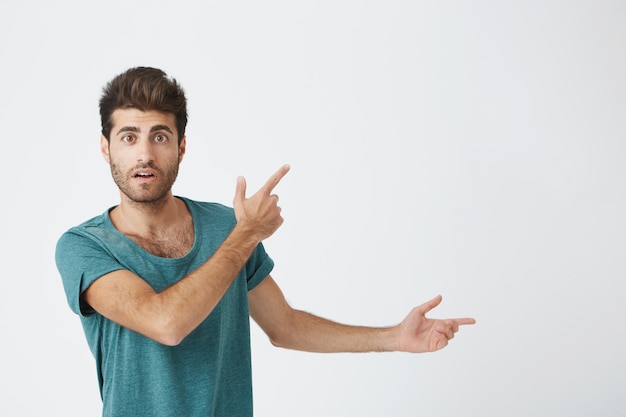 Innenaufnahme eines erstaunten gutaussehenden Mannes in lässigem Stoff, der einen faszinierten Blick erregt hat und mit seinem Zeigefinger auf eine leere Wand hinweist. Isolierter Schuss, horizontal