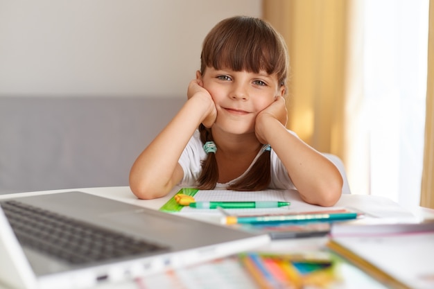 Innenaufnahme eines glücklichen positiven weiblichen Kindes, das mit optimistischem Gesichtsausdruck in die Kamera schaut, Hausaufgaben macht, mag Online-Unterricht während der Quarantäne.