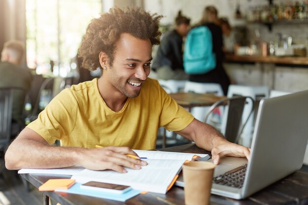 Innenaufnahme eines glücklichen Studentenmännchens mit lockigem Haar, das beiläufig gekleidet in der Cafeteria gekleidet arbeitet, die mit modernen Technologien arbeitet, während das Lernen mit einem Lächeln im Notizbuch studiert, das Nachricht von Freund empfängt